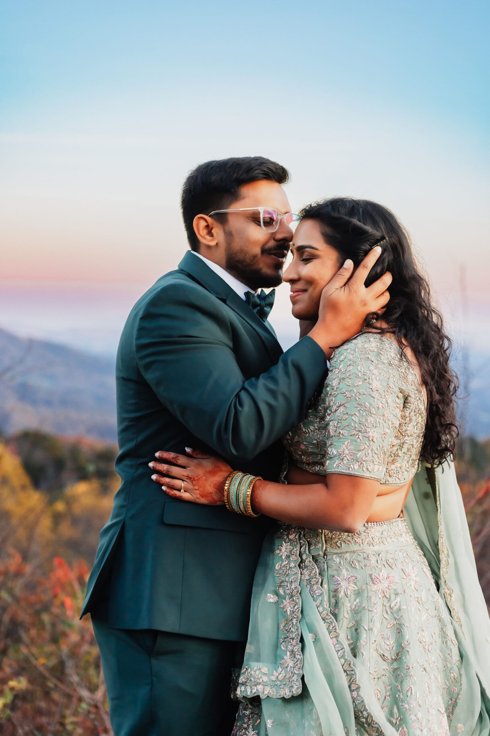 Indian couple in a in green wedding tux and lehenga in a field of green grass with a rust colored mountain in the background with blue rolling mountains in the background. They are standing close and kissing.