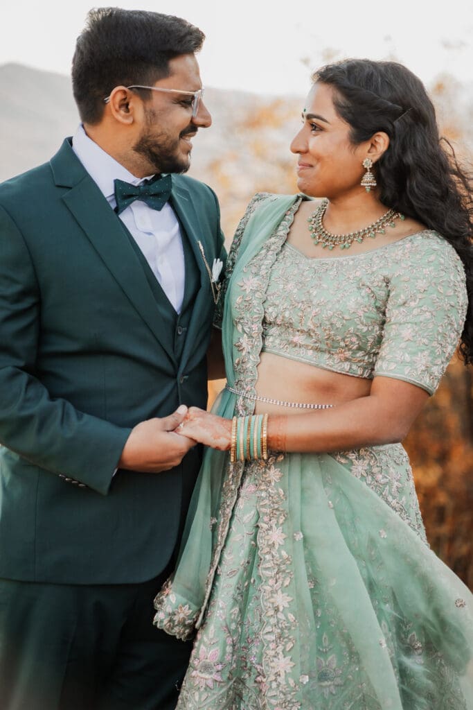 Indian couple in a in green wedding tux and lehenga in a field of green grass with a rust colored mountain in the background with blue rolling mountains in the background. They are standing close and kissing.