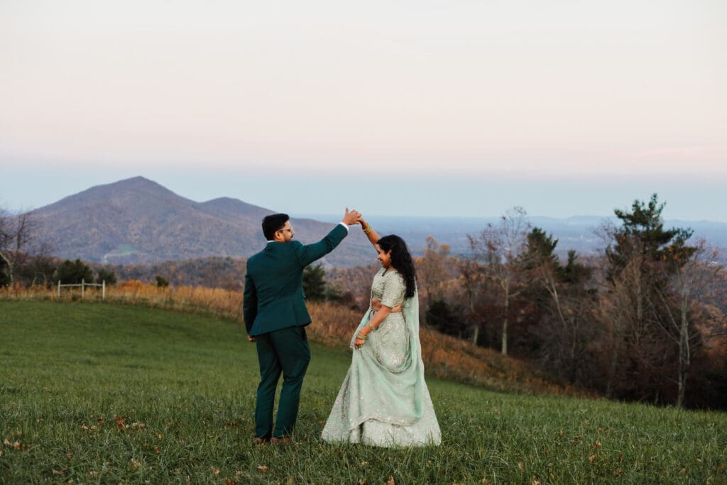Indian couple in a in green wedding tux and lehenga in a field of green grass with a rust colored mountain in the background with blue rolling mountains in the background. They are doing the dancing in the field.