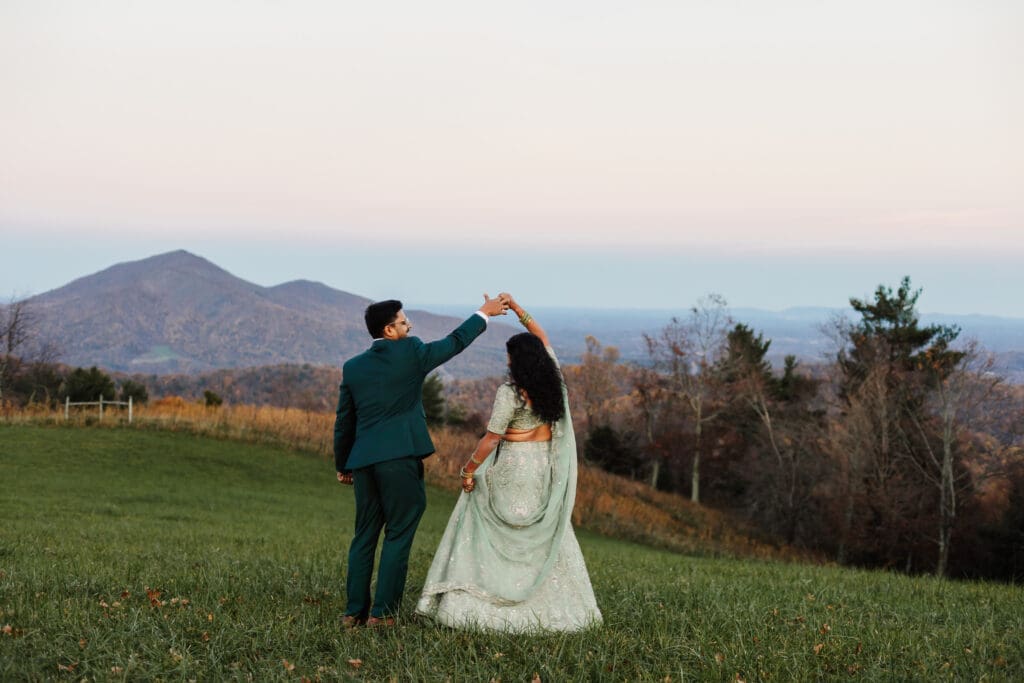 Indian couple in a in green wedding tux and lehenga in a field of green grass with a rust colored mountain in the background with blue rolling mountains in the background. They are doing the dancing in the field.