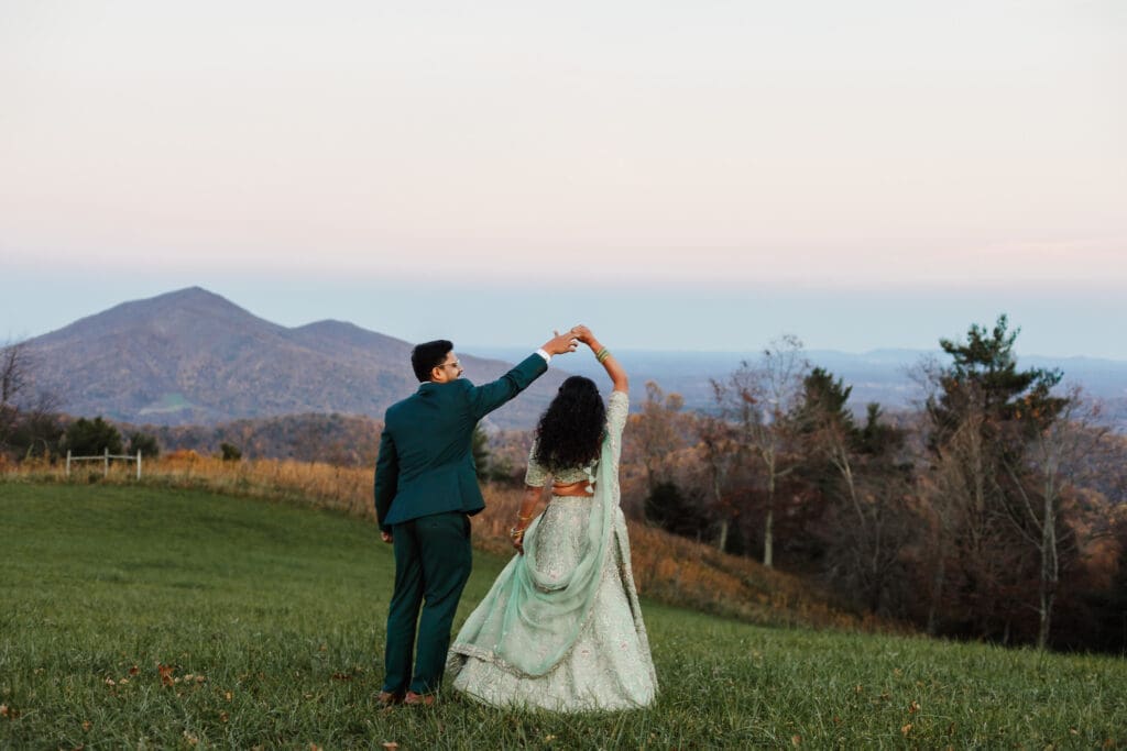 Indian couple in a in green wedding tux and lehenga in a field of green grass with a rust colored mountain in the background with blue rolling mountains in the background. They are doing the dancing in the field.
