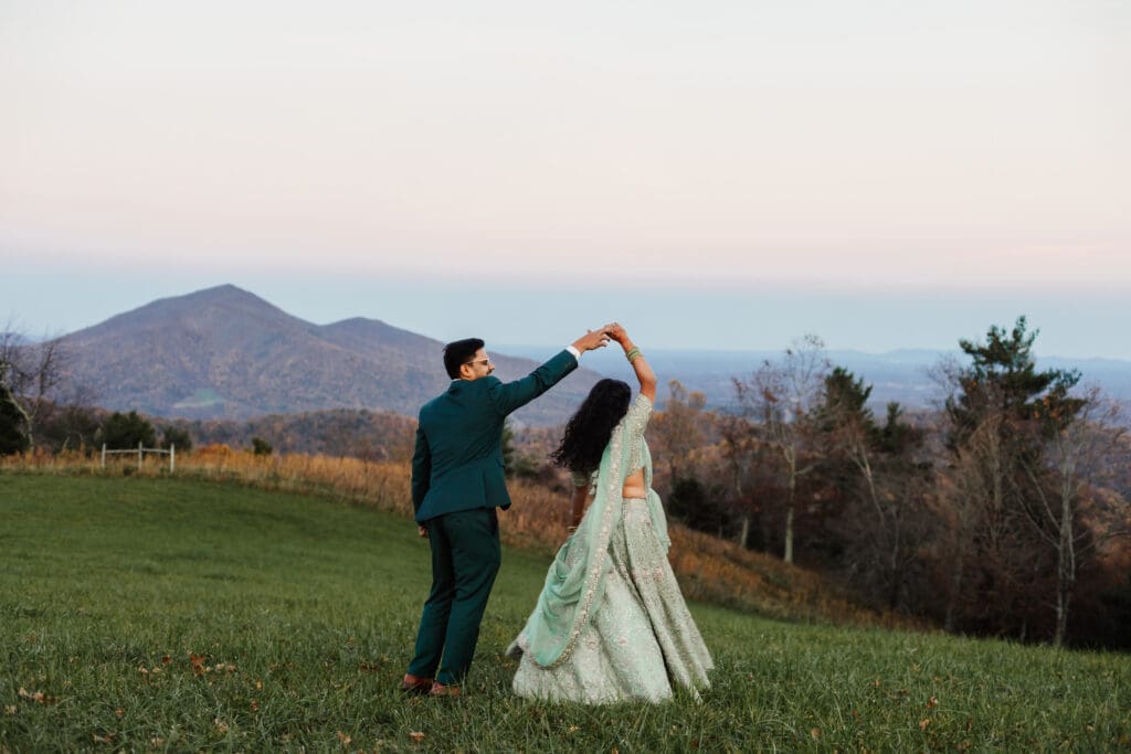 Indian couple in a in green wedding tux and lehenga in a field of green grass with a rust colored mountain in the background with blue rolling mountains in the background. They are doing the dancing in the field.