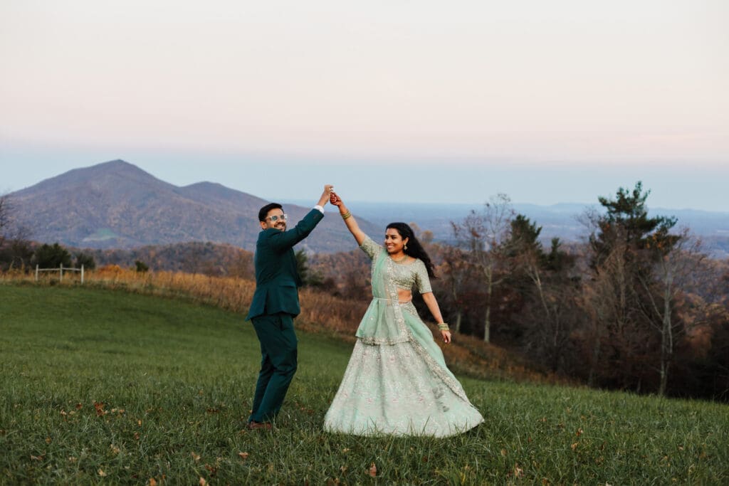 Indian couple in a in green wedding tux and lehenga in a field of green grass with a rust colored mountain in the background with blue rolling mountains in the background. They are doing the dancing in the field.