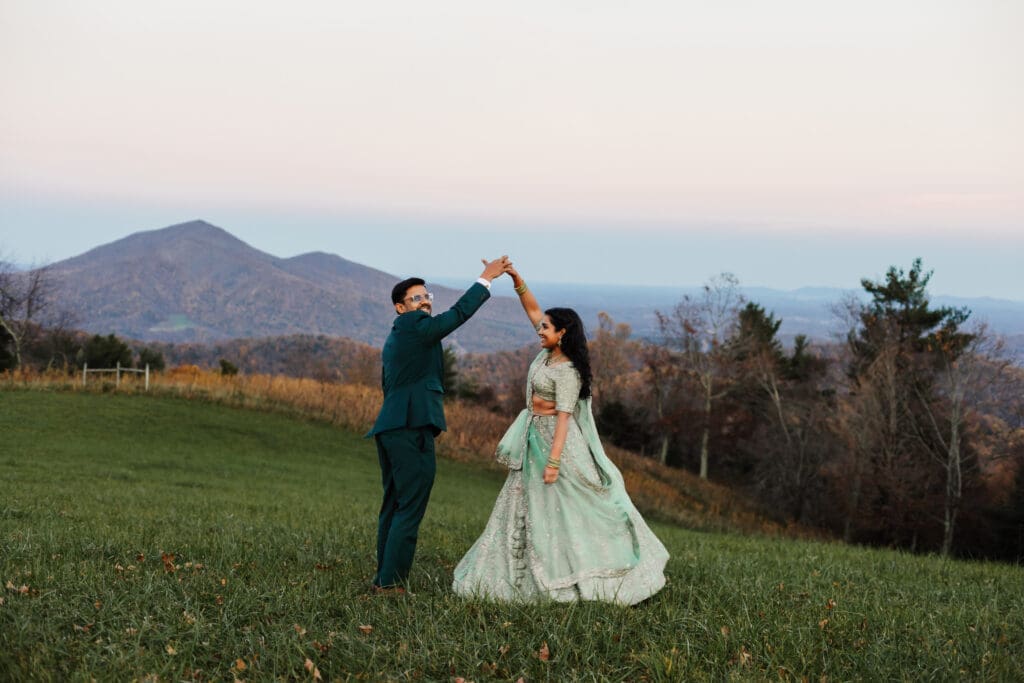 Indian couple in a in green wedding tux and lehenga in a field of green grass with a rust colored mountain in the background with blue rolling mountains in the background. They are doing the dancing in the field.