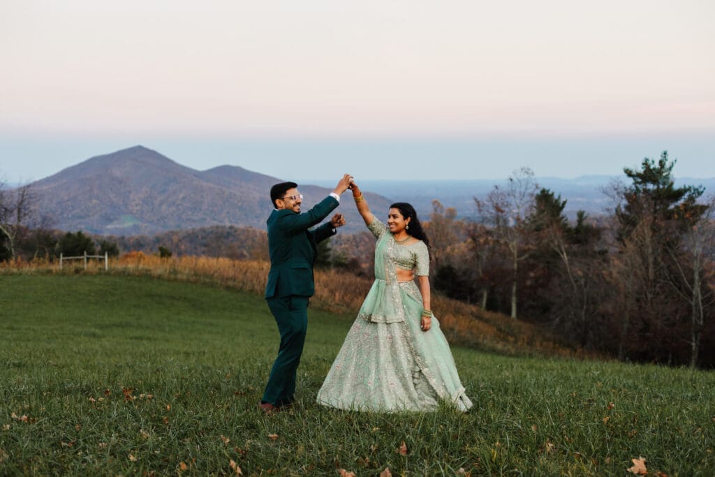 Indian couple in a in green wedding tux and lehenga in a field of green grass with a rust colored mountain in the background with blue rolling mountains in the background. They are doing the dancing in the field.