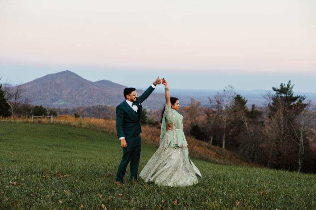 Indian couple in a in green wedding tux and lehenga in a field of green grass with a rust colored mountain in the background with blue rolling mountains in the background. They are doing the dancing in the field.