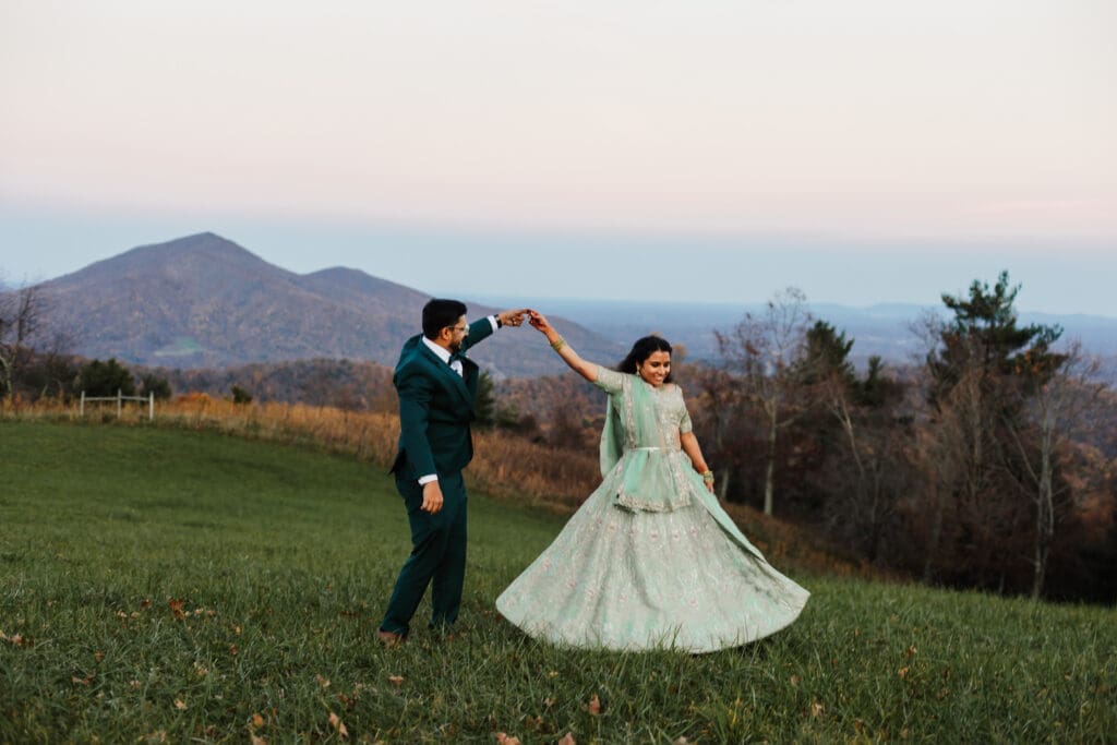 Indian couple in a in green wedding tux and lehenga in a field of green grass with a rust colored mountain in the background with blue rolling mountains in the background. They are doing the dancing in the field.