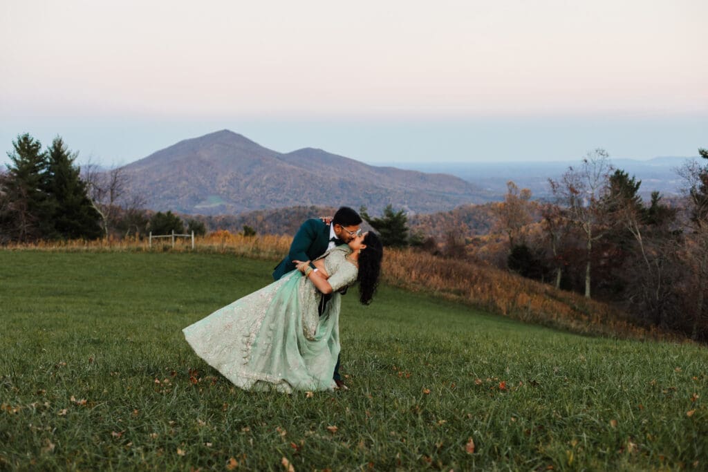 Indian couple in a in green wedding tux and lehenga in a field of green grass with a rust colored mountain in the background with blue rolling mountains in the background. They are doing the dip kiss pose.