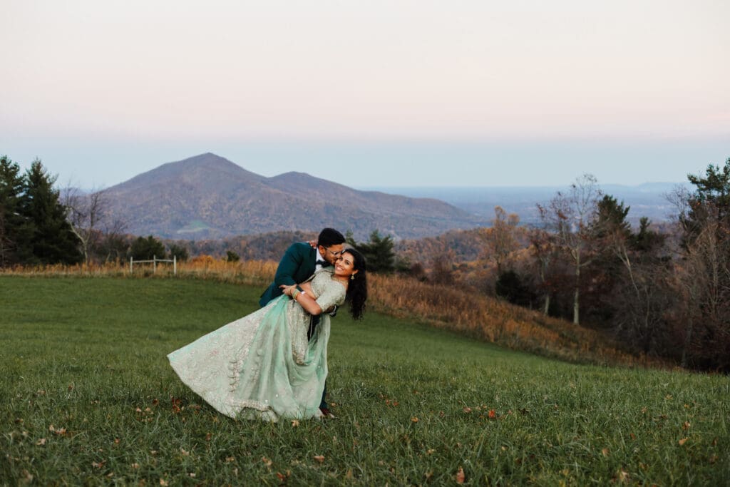 Indian couple in a in green wedding tux and lehenga in a field of green grass with a rust colored mountain in the background with blue rolling mountains in the background. They are doing the dip kiss pose.