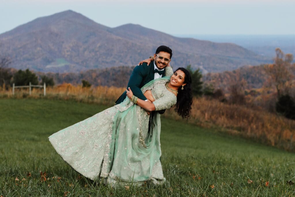 Indian couple in a in green wedding tux and lehenga in a field of green grass with a rust colored mountain in the background with blue rolling mountains in the background. They are doing the dip kiss pose.