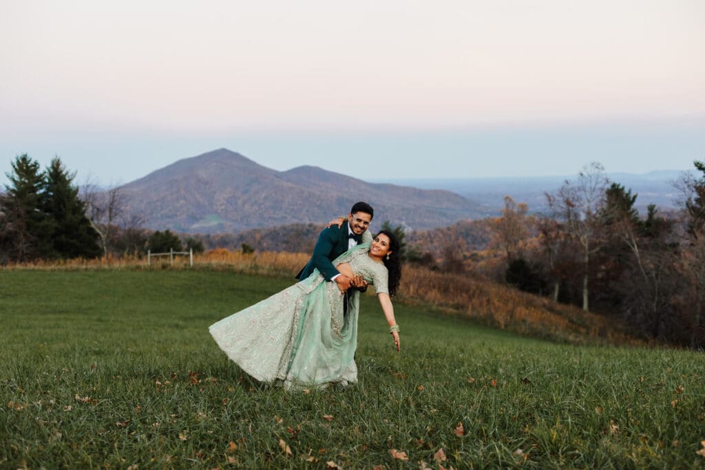 Indian couple in a in green wedding tux and lehenga in a field of green grass with a rust colored mountain in the background with blue rolling mountains in the background. They are doing the dip kiss pose.