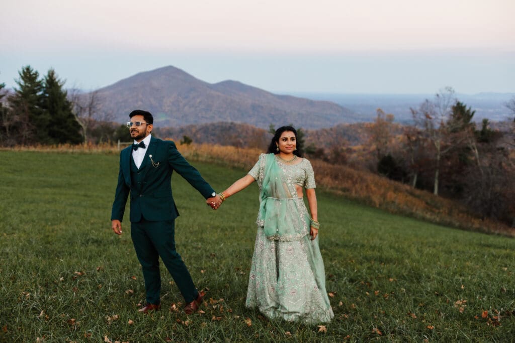 Indian couple in a in green wedding tux and lehenga in a field of green grass with a rust colored mountain in the background with blue rolling mountains in the background. They are holding hands while staring off into the distance.