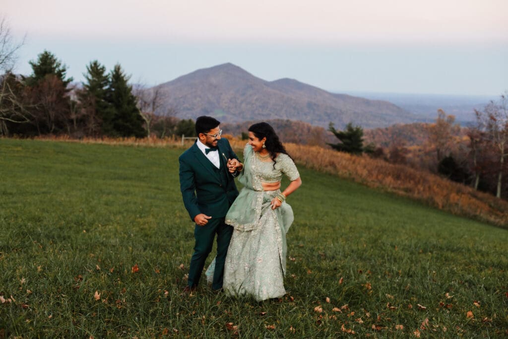 Indian couple in a in green wedding tux and lehenga in a field of green grass with a rust colored mountain in the background with blue rolling mountains in the background. They are frolicking in the field looking at each other.