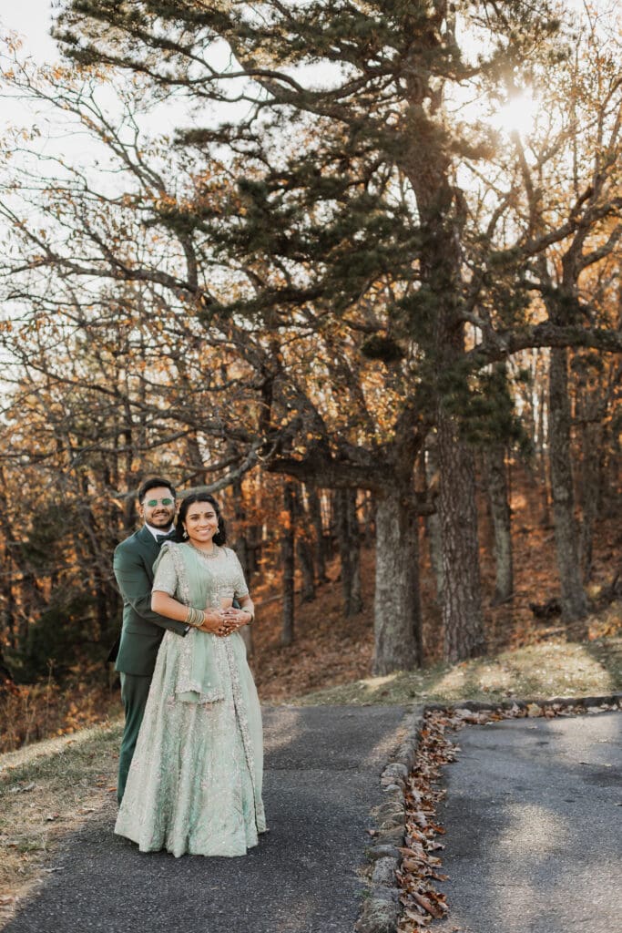 Indian couple in a in green wedding tux and lehenga in a field of green grass with a rust colored mountain in the background with blue rolling mountains in the background. They are standing close and kissing.