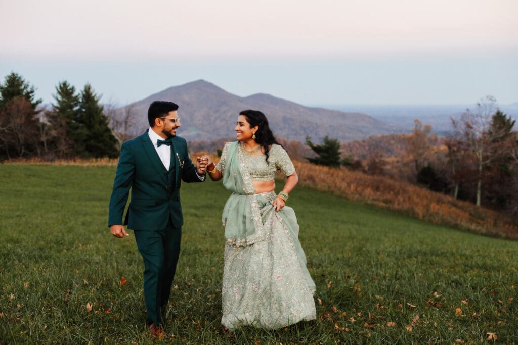 Indian couple in a in green wedding tux and lehenga in a field of green grass with a rust colored mountain in the background with blue rolling mountains in the background. They are frolicking in the field looking at each other.