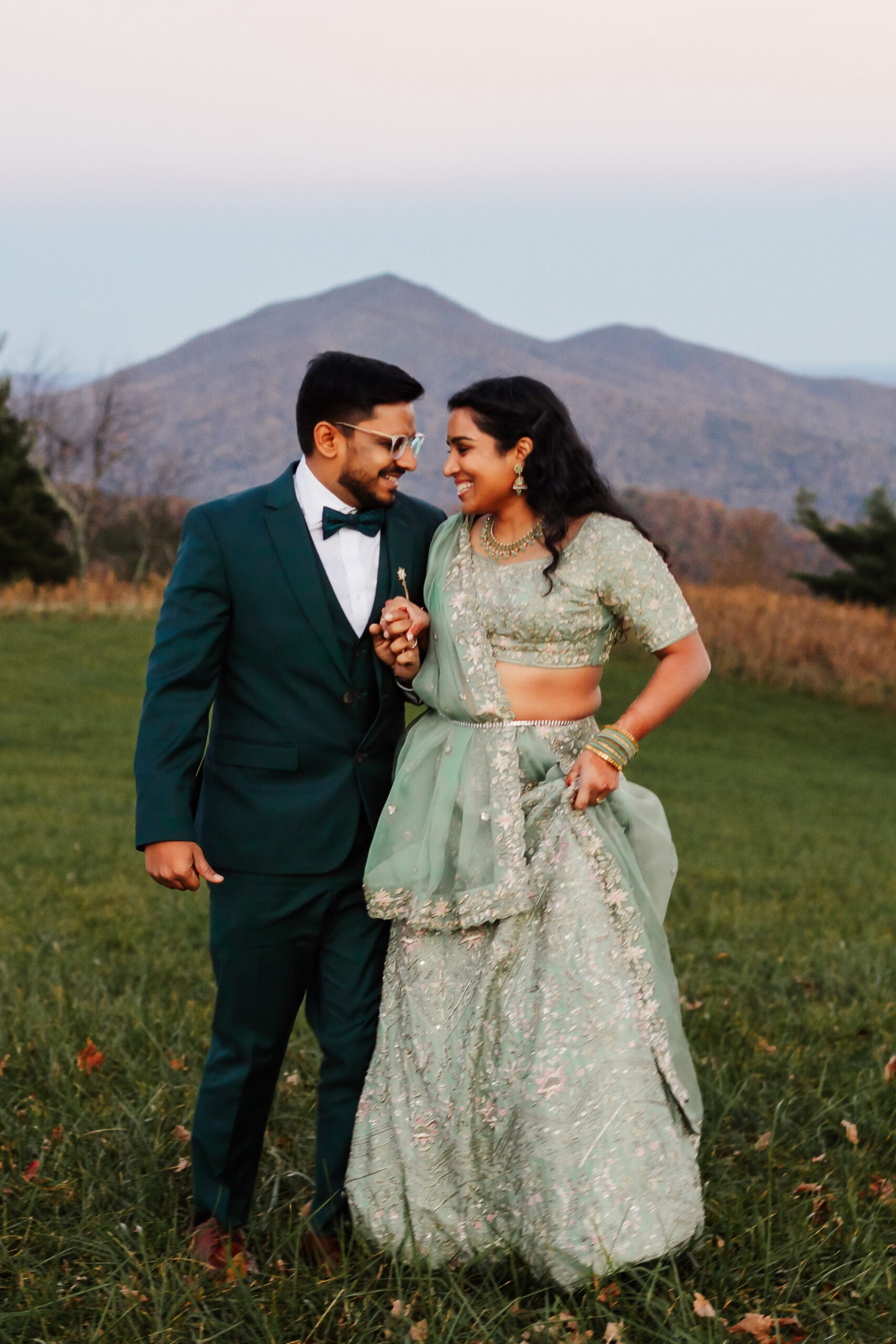 Indian couple in a in green wedding tux and lehenga in a field of green grass with a rust colored mountain in the background with blue rolling mountains in the background. They are frolicking in the field looking at each other.