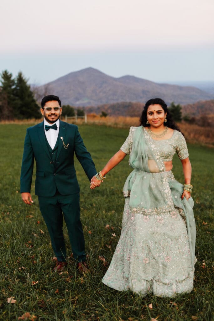Indian couple in a in green wedding tux and lehenga in a field of green grass with a rust colored mountain in the background with blue rolling mountains in the background. They are frolicking in the field looking at each other.