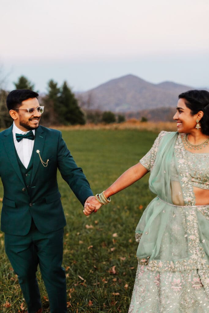 Indian couple in a in green wedding tux and lehenga in a field of green grass with a rust colored mountain in the background with blue rolling mountains in the background. They are frolicking in the field looking at each other.