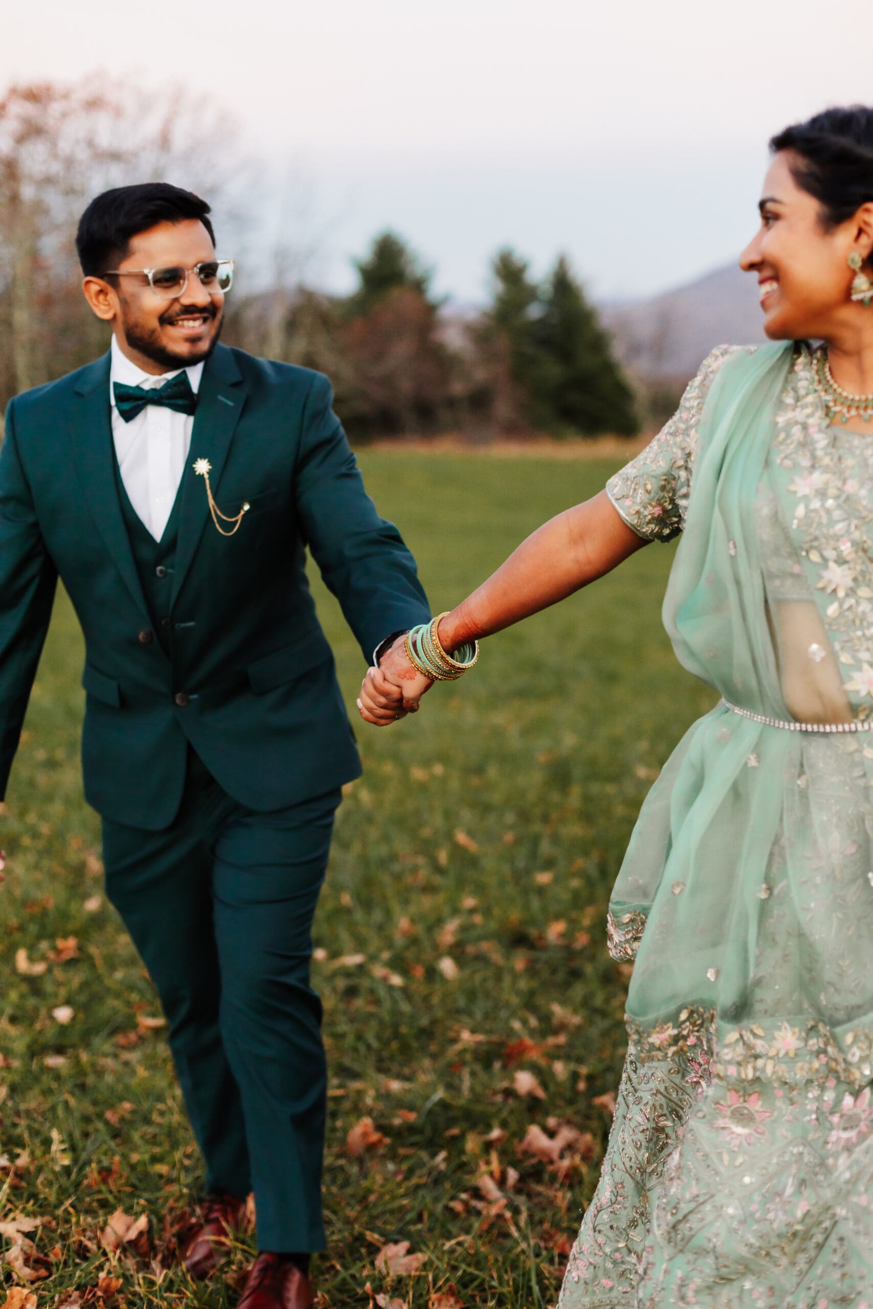 Indian couple in a in green wedding tux and lehenga in a field of green grass with a rust colored mountain in the background with blue rolling mountains in the background. They are frolicking in the field looking at each other.