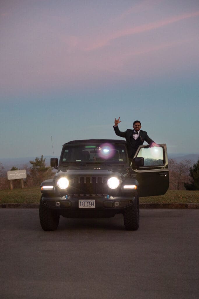 Indian couple in a in green wedding tux and lehenga in a field of green grass with a rust colored mountain in the background with blue rolling mountains in the background. They are in the dark being illuminated by the jeep lights and dancing.