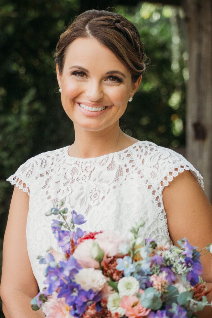 Bride at elopement standing in front of woods smiling big while holding her bouquet.