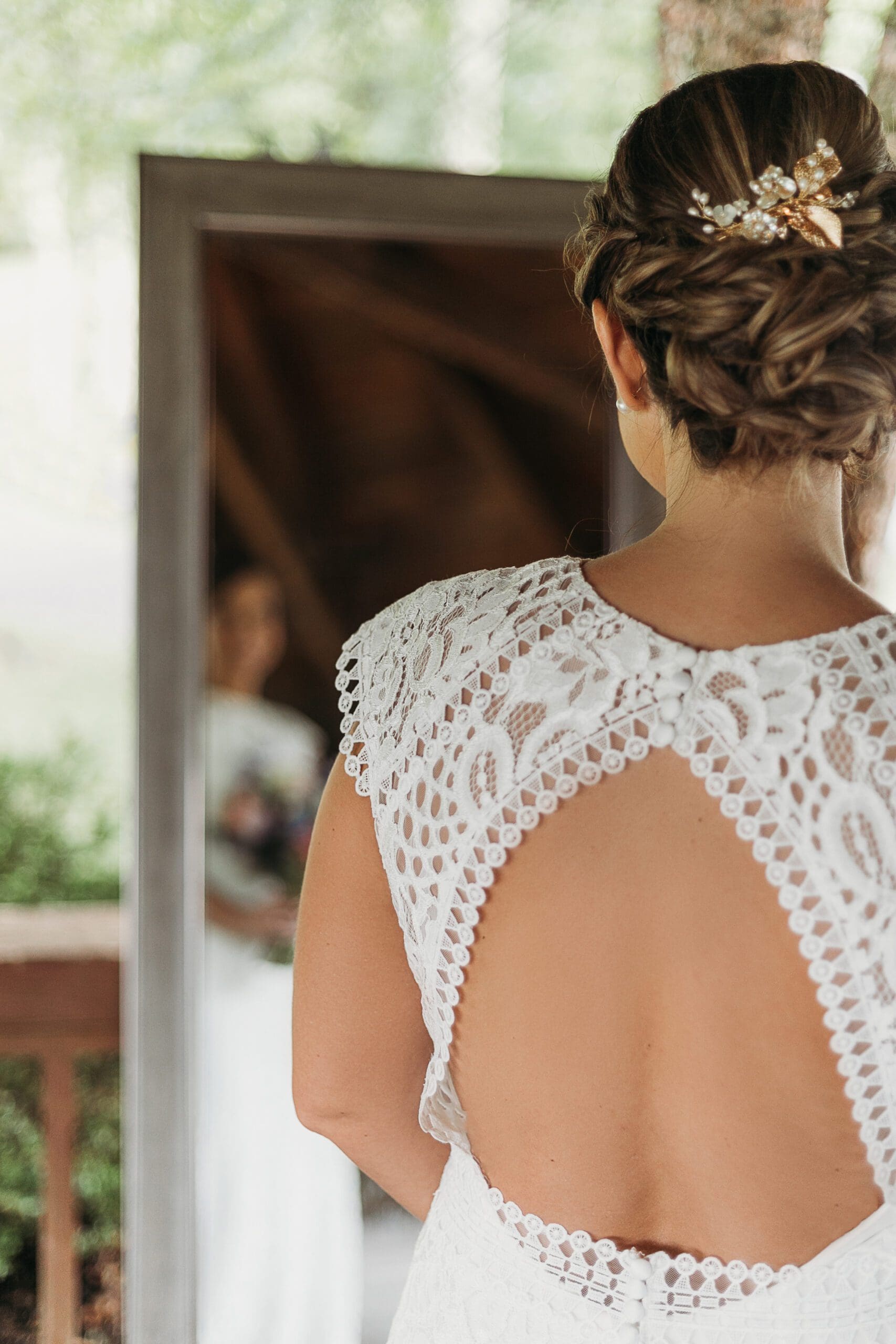 Bride standing in front of a mirror looking at herself with the back of her dress in focus. The mirror is under a gazebo surrounded by greenery.