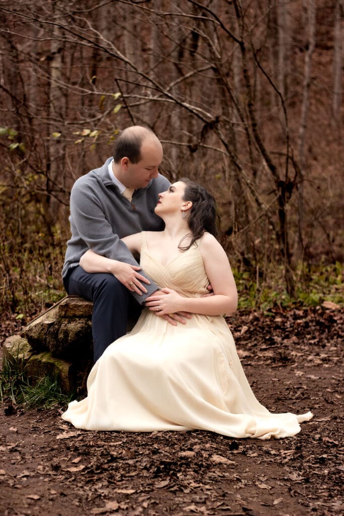 Couple in woods sitting on ground and logs with wedding attire on gazing into each others eyes with arms embracing.