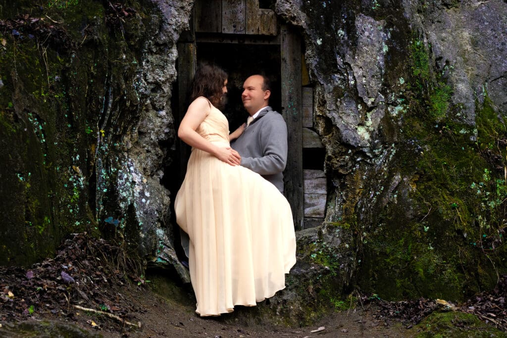 Couple sitting ion cave ledge in winter with their wedding attire on a few spots of greenery popping out in a sea of brown.