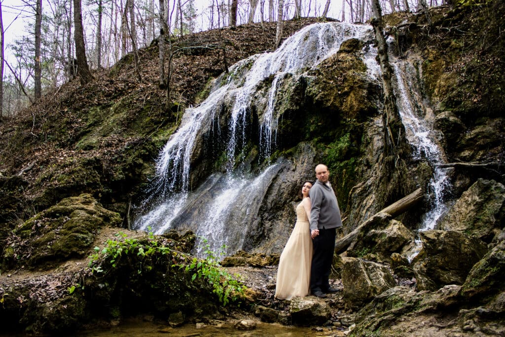 Couple standing in front of a waterfall in winter with their wedding attire on a few spots of greenery popping out in a sea of brown. This is a waterfall elopement