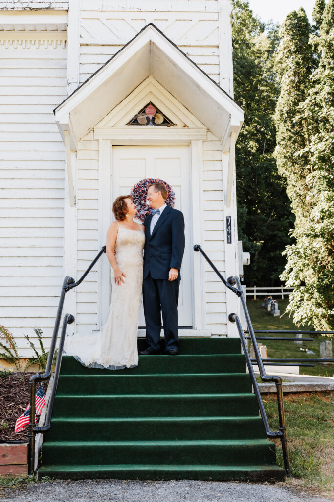 Couple eloping at a small chapel in the blue ridge mountains standing outside the white chapel with lush green landscape.