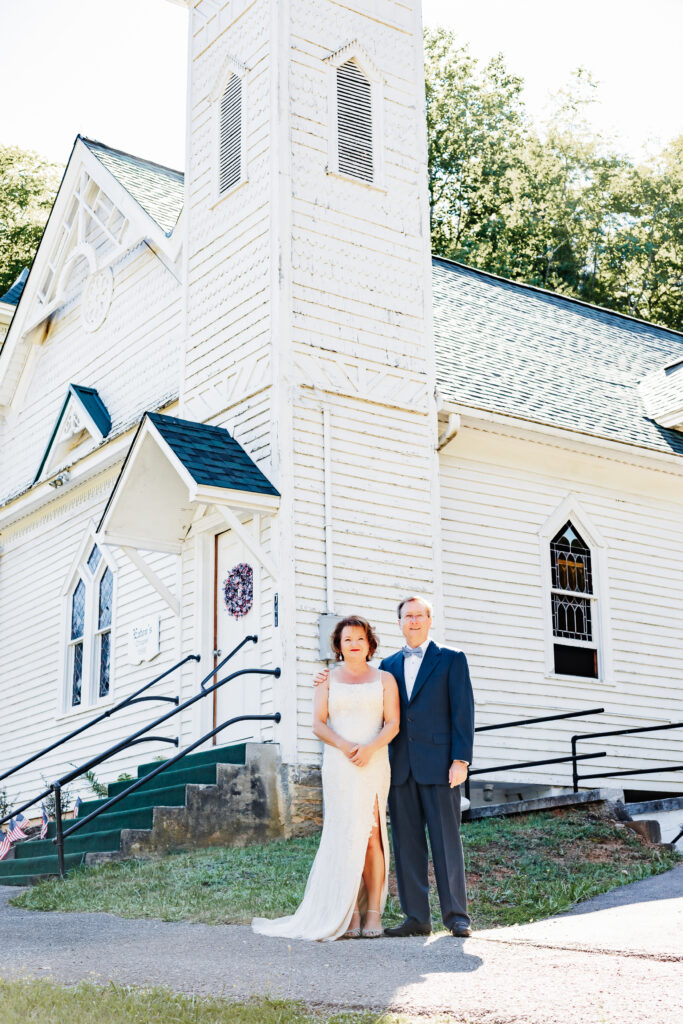 Couple eloping at a small chapel in the blue ridge mountains standing outside the white chapel with lush green landscape.