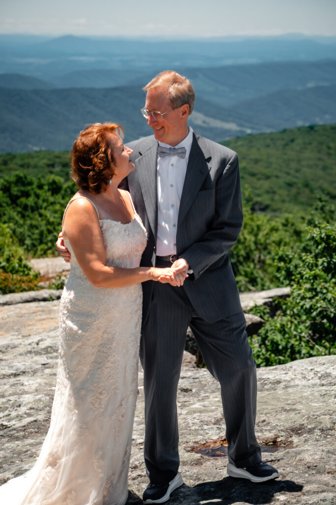 Couple standing on mountain ledge eloping with green rolling mountains in the background and blue skies as far as the eyes can see. She has a white dress and he has a grey suit with a blue bow tie.