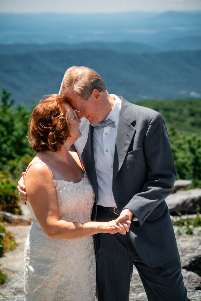 Couple standing on mountain ledge eloping with green rolling mountains in the background and blue skies as far as the eyes can see. She has a white dress and he has a grey suit with a blue bow tie.