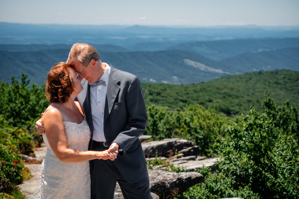 Couple standing on mountain ledge eloping with green rolling mountains in the background and blue skies as far as the eyes can see. She has a white dress and he has a grey suit with a blue bow tie.
