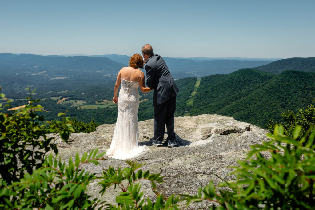 Couple standing on mountain ledge eloping with green rolling mountains in the background and blue skies as far as the eyes can see. She has a white dress and he has a grey suit with a blue bow tie.