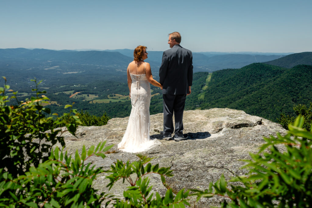 Couple standing on mountain ledge eloping with green rolling mountains in the background and blue skies as far as the eyes can see. She has a white dress and he has a grey suit with a blue bow tie.