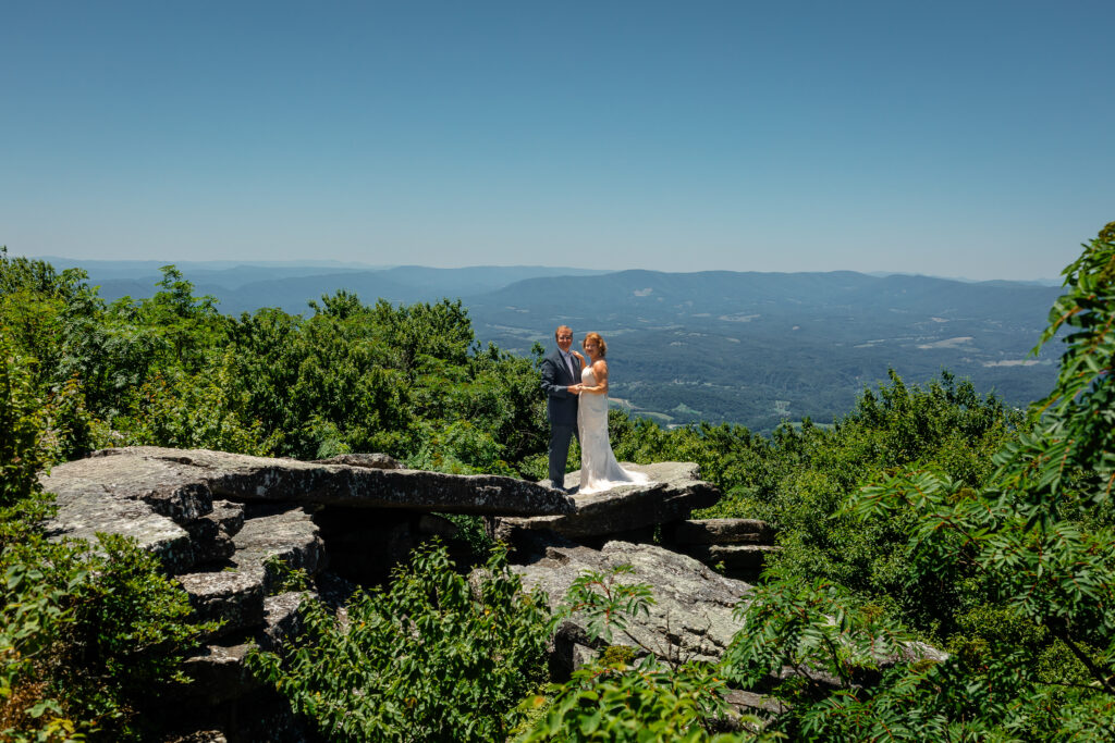Couple standing on mountain ledge eloping with green rolling mountains in the background and blue skies as far as the eyes can see. She has a white dress and he has a grey suit with a blue bow tie.