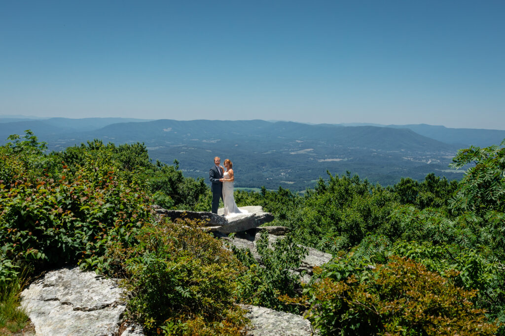 Couple standing on mountain ledge eloping with green rolling mountains in the background and blue skies as far as the eyes can see. She has a white dress and he has a grey suit with a blue bow tie.