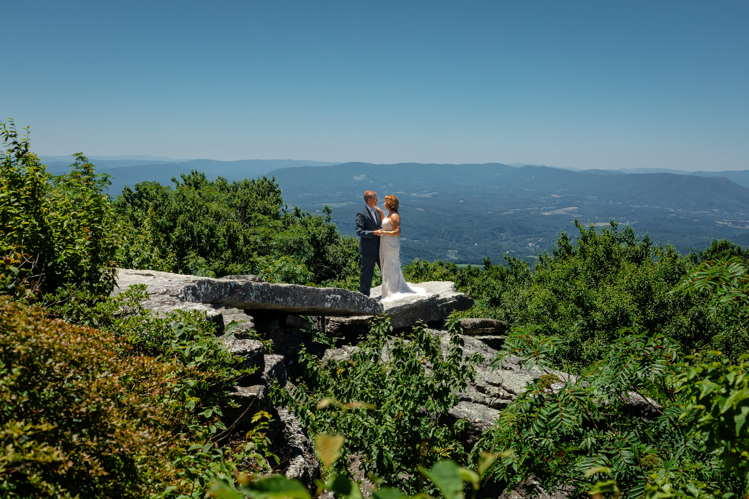 Couple standing on mountain ledge eloping with green rolling mountains in the background and blue skies as far as the eyes can see. She has a white dress and he has a grey suit with a blue bow tie.