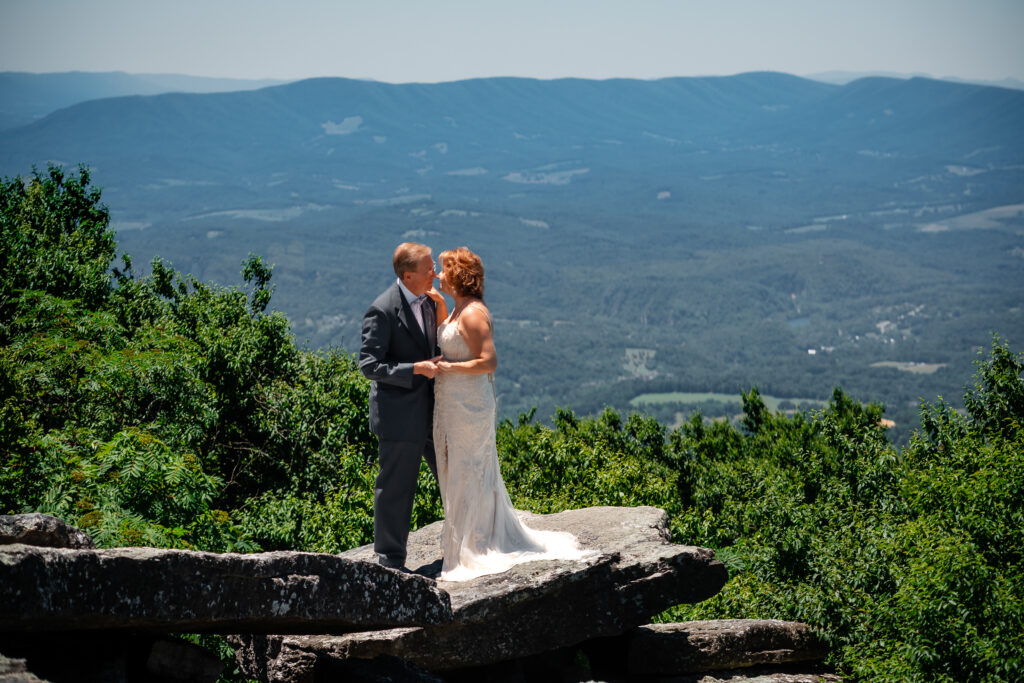 Couple standing on mountain ledge eloping with green rolling mountains in the background and blue skies as far as the eyes can see. She has a white dress and he has a grey suit with a blue bow tie.