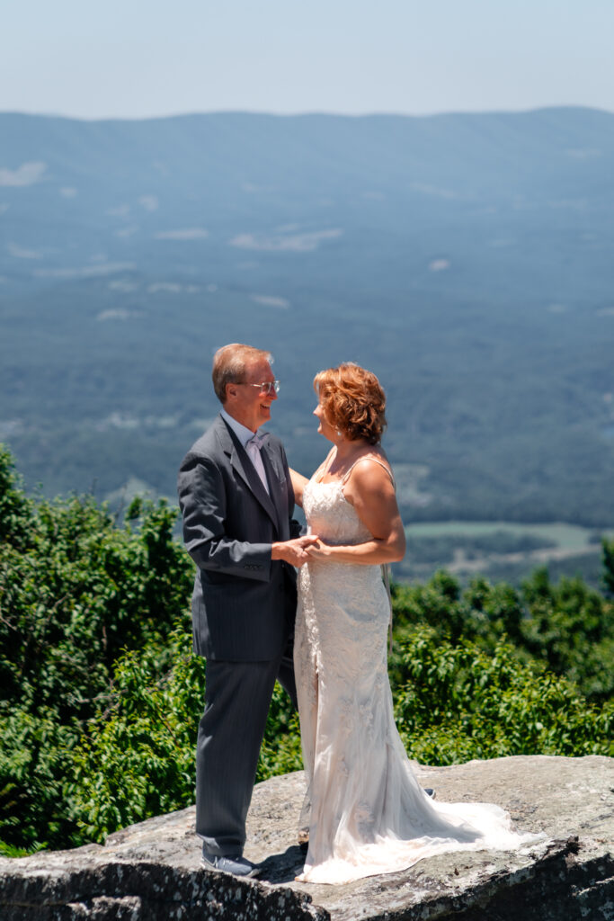 Couple standing on mountain ledge eloping with green rolling mountains in the background and blue skies as far as the eyes can see. She has a white dress and he has a grey suit with a blue bow tie.