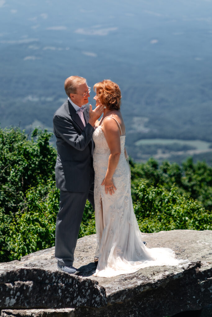Couple standing on mountain ledge eloping with green rolling mountains in the background and blue skies as far as the eyes can see. She has a white dress and he has a grey suit with a blue bow tie.