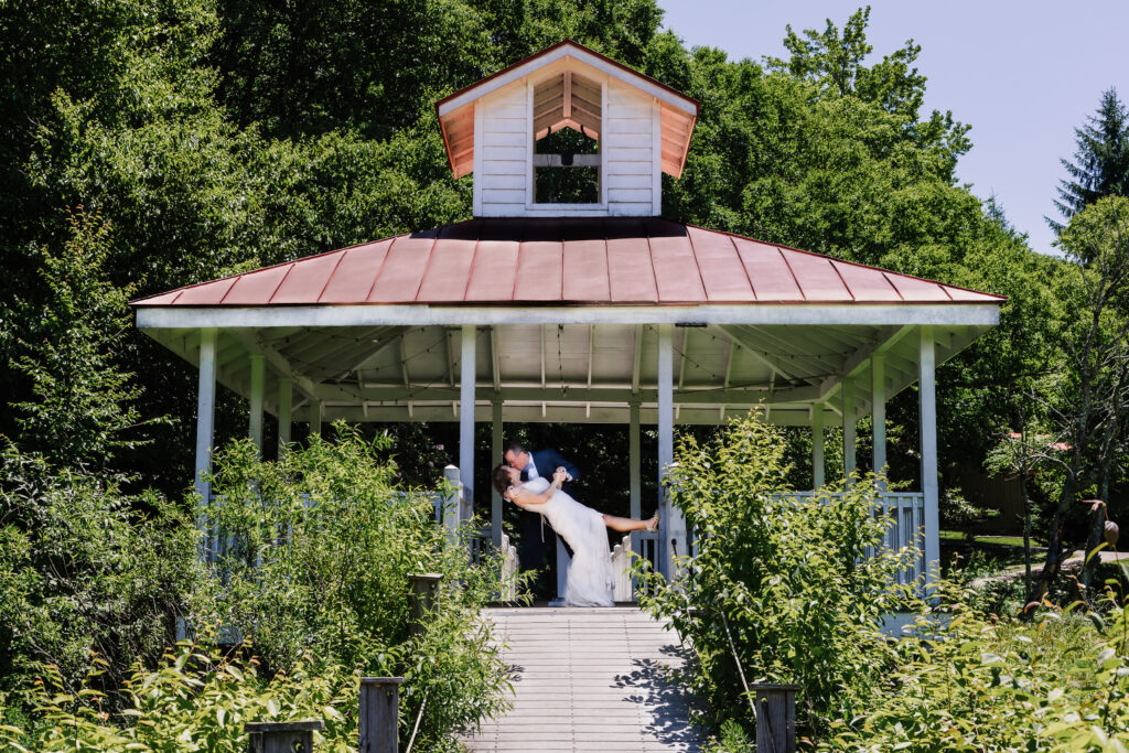 Couple in gazebo with white wood and a red roof. They are in wedding clothes from their elopement. There is greenery and shrubs growing up around the pier walkway.