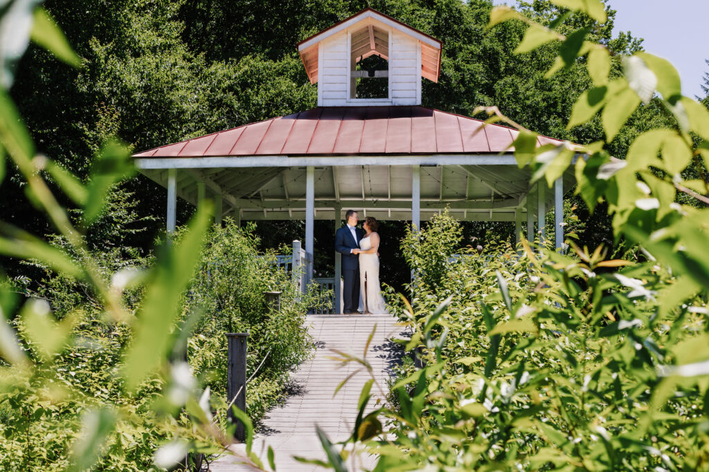 Couple in gazebo with white wood and a red roof. They are in wedding clothes from their elopement. There is greenery and shrubs growing up around the pier walkway.