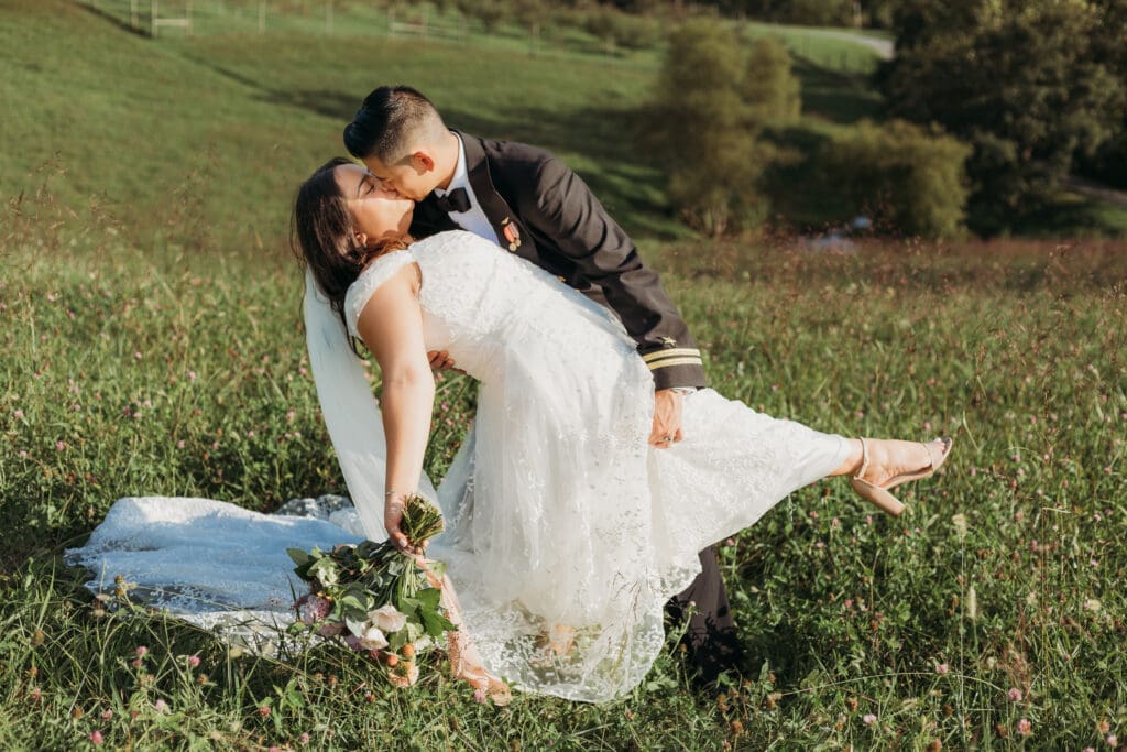 Couple in wedding attire eloping in a field of flowers surrounded by greenery. He is dip kissing her.