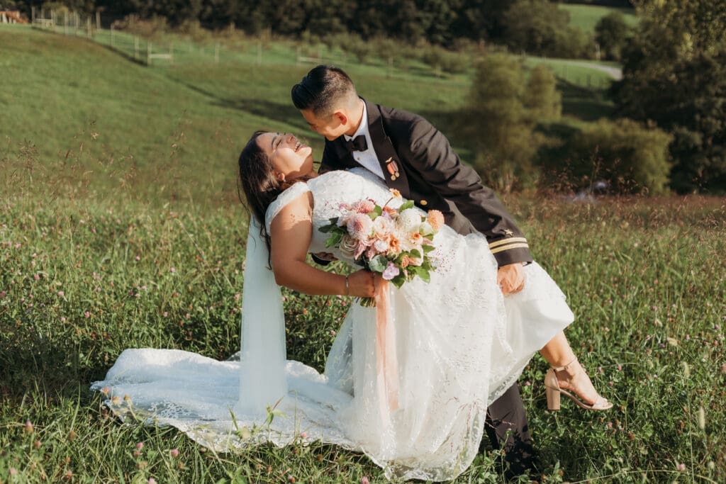 Couple in wedding attire eloping in a field of flowers surrounded by greenery. He is dip kissing her.