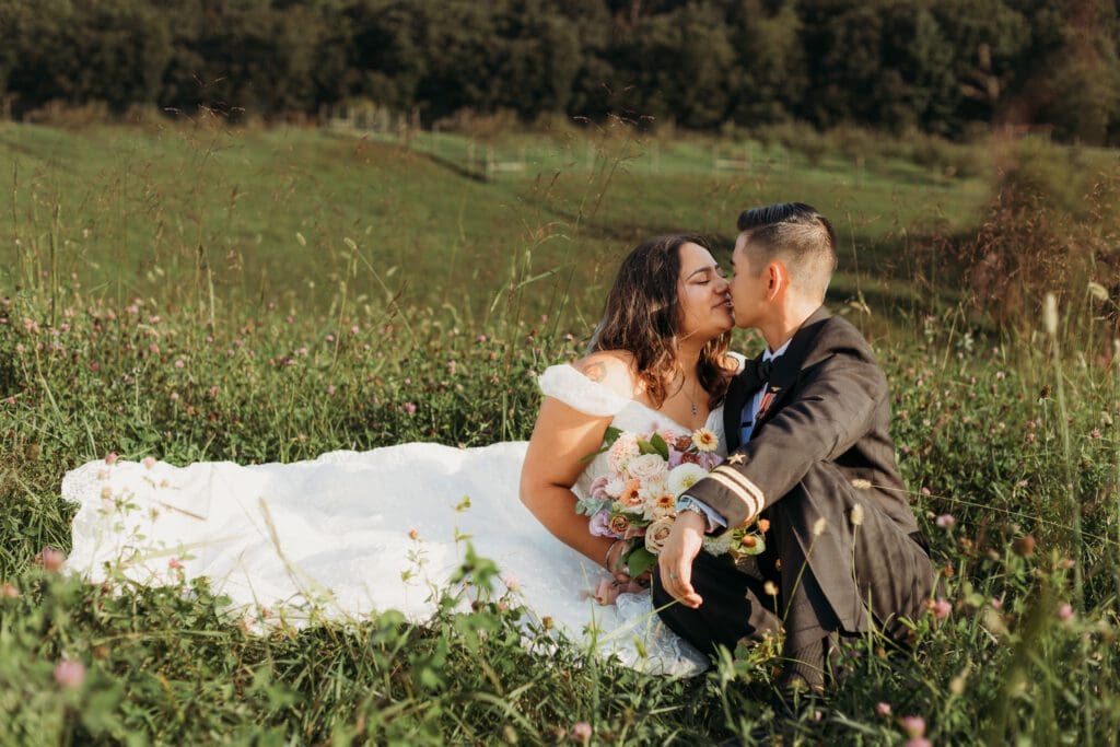 Couple in wedding attire eloping in a field of flowers surrounded by greenery. They are sitting in the grass and flowers kissing.