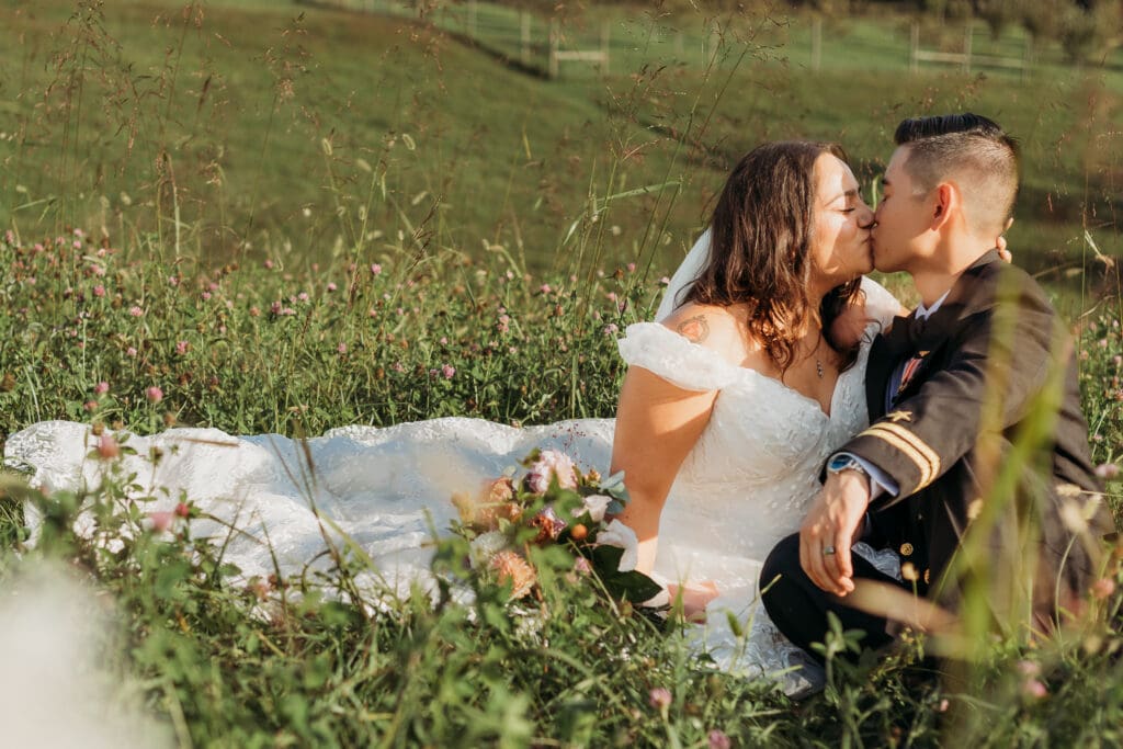 Couple in wedding attire eloping in a field of flowers surrounded by greenery. They are sitting in the grass and flowers kissing.