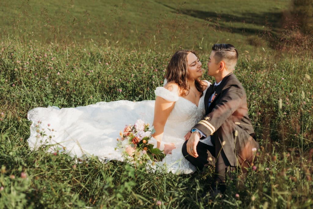 Couple in wedding attire eloping in a field of flowers surrounded by greenery. They are sitting in the grass and flowers kissing.