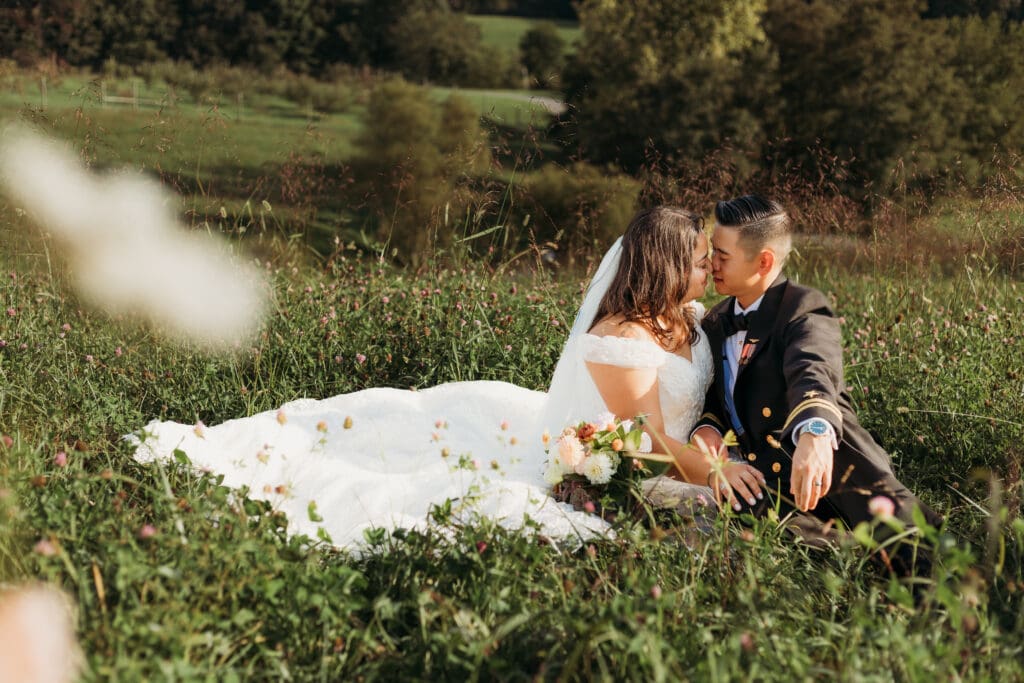 Couple in wedding attire eloping in a field of flowers surrounded by greenery. They are sitting in the grass and flowers kissing.